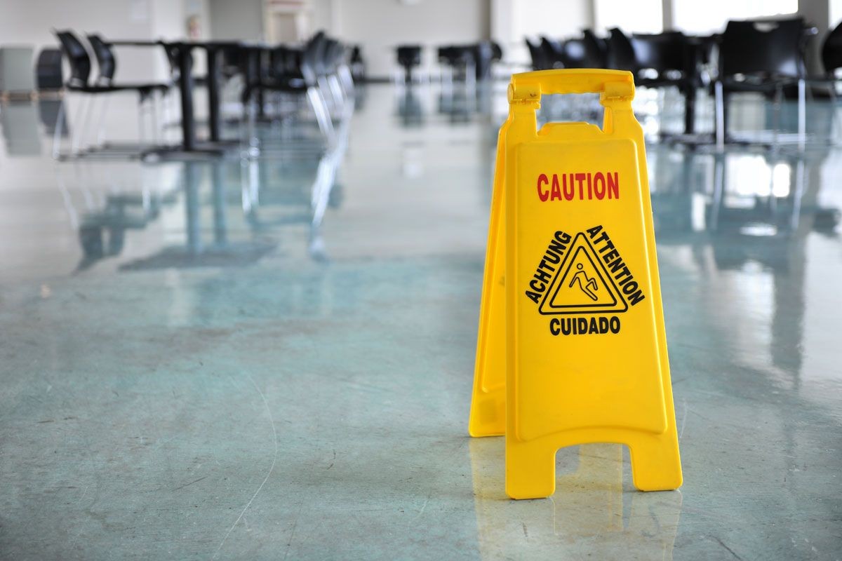 Yellow caution sign on a polished floor in a cafeteria, warning in multiple languages about slippery conditions.